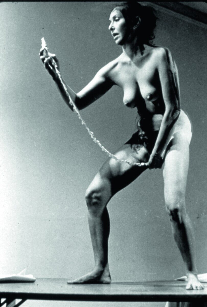 Black-and-white photo of Carolee Schneemann pulling a written manifesto out of her vagina in her 1975 performance piece