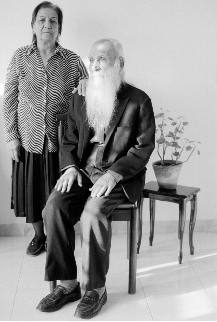 An older man and woman, the former sitting, the latter standing, with a potted plant