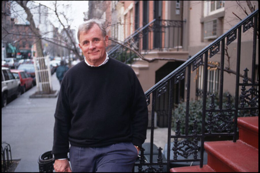 A portrait of Edmund White wearing a navy-blue sweater and standing on a red-staired NYC stoop with a wrought-iron railing